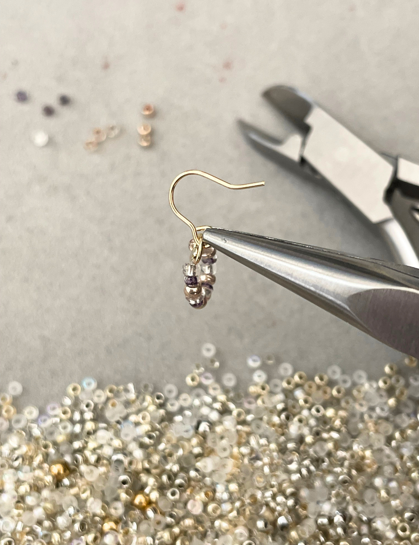 Gold earring with beads being adjusted with pliers on a gray surface with beads scattered around.